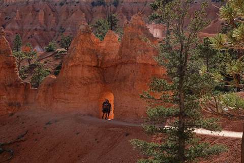 Two hikers pass through a hole carved in limestone feature along a trail lined with trees and rocks