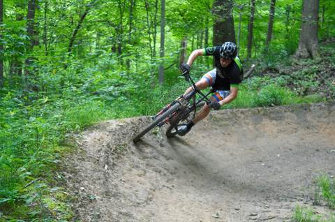 A mountain biker wearing a helmet banks around a curve at a 45-degree angle, on a wooded trail.
