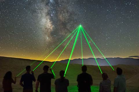 Seven people silhouetted pointing green lasers to the same spot in a dark night sky.