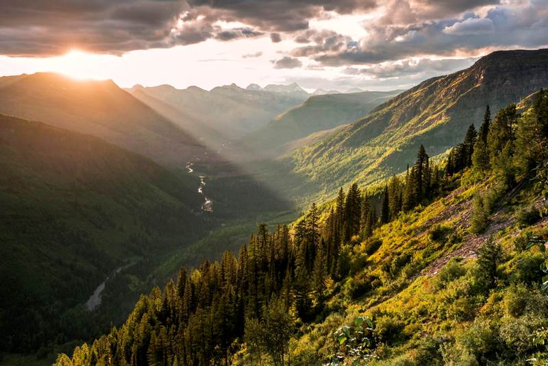 The sun sets atop a ridge of mountains; a stream glistens in the mountain valley below.