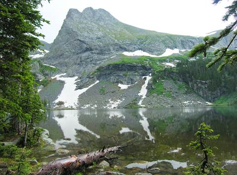 An alpine lake with conifer trees at the side, and rocky summit with snow