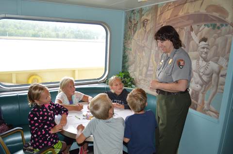 Children sit around a table working on Junior Ranger booklets while a Park Ranger stands nearby.