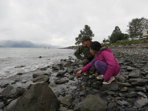 A park ranger and a child standing on a rocky beach. They are looking out into the water