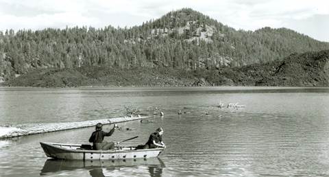 Two women fishing from a row boat on a lake backed by a lava flow and a volcanic peak.